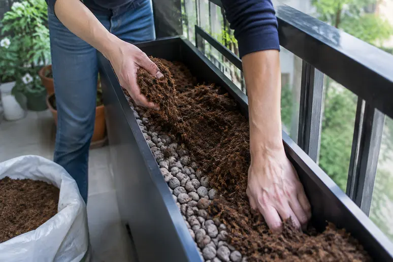Hände beim Befüllen eines schmalen Hochbeets mit Pflanzenerde auf dem Balkon